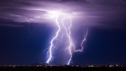 Nachtaufnahme eines Gewitters über einer Stadt: Mehrere verzweigte, hell leuchtende Blitze durchziehen den violett-dunkelblauen Himmel und schlagen in einen Hügel und die Landschaft ein. Am Horizont sind zahlreiche kleine Lichter der Stadt zu erkennen.
