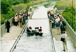 In einem etwa fünf Meter breiten Kanal steht eine einzelne Welle vor einer Gruppe in einem Motorboot. Am Rand stehen Schaulustige.