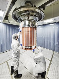 Zwei Wissenschaftler im Schutzanzug arbeiten an einem von der Decke hängenden, kupferfarbenen Zylinder.