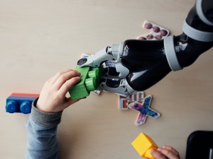 Zu sehen ist eine Kinderhand und eine Roboterhand, die gemeinsam einen Spielzeugbauklotz in die Luft halten. Im Hintergrund ist ein Tisch und weiteres Spielzeug zu sehen. 
