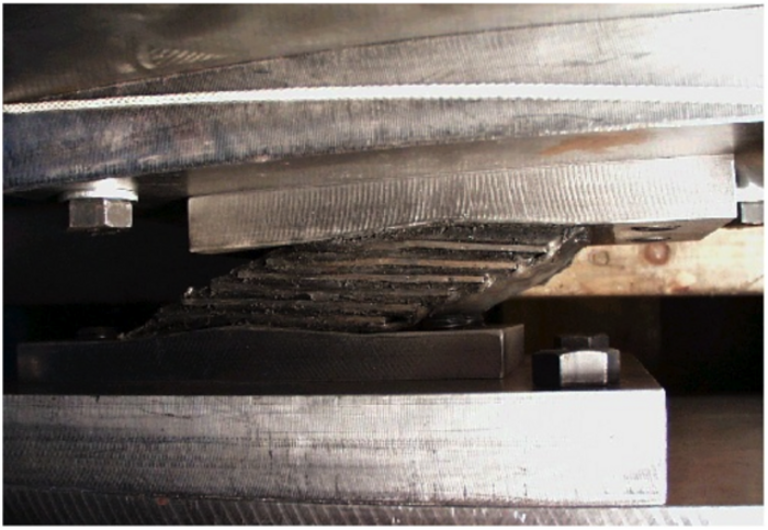 Basisisolierung Zwei massive Metallplatten, dazwischen ein prismenförmiges Lager, das aus dünneren Platten besteht. Diese sind mit einer harzartigen Masse verbunden. Das Lager hat einen parallelogrammförmigen Querschnitt.