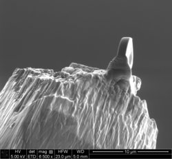 Eine winzige Röntgenlinse sitzt auf einer Nadelspitze. Unter dem Elektronenmikroskop wirkt sie wie ein Fels auf einem schroffen Bergkamm.