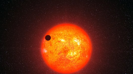 Großer sonnenähnlicher Stern, links oben deckt ein kleines schwarzes kreisförmiges Objekt einen Teil des Sterns ab.