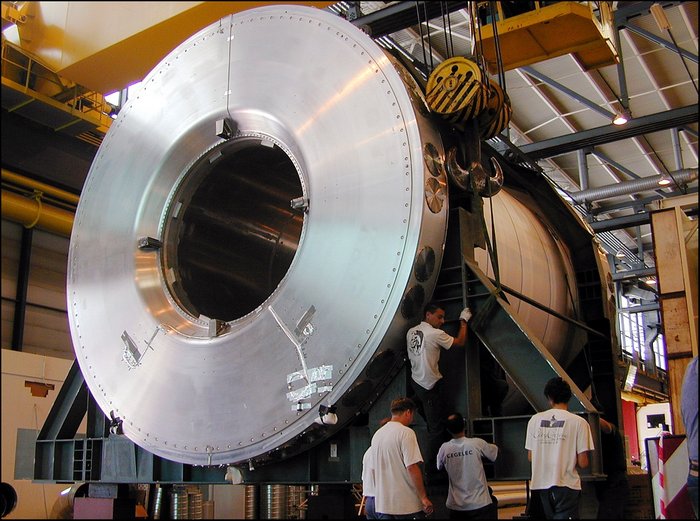 Der Kryostat für das Kalorimeter des ATLAS-Detektors Foto von fünf Männern vor einer riesigen metallenen Röhre, die in einer Montagehalle an einem Kran hängt.