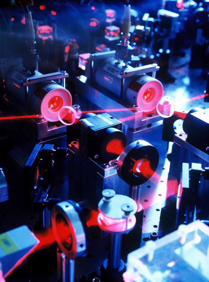 Aufbau der Laserapparatur Ein komplexer Aufbau aus Spiegeln, Linsen und Laserköpfen. Der Laserstrahl ist in unterschiedlicher Dicke erkennbar.