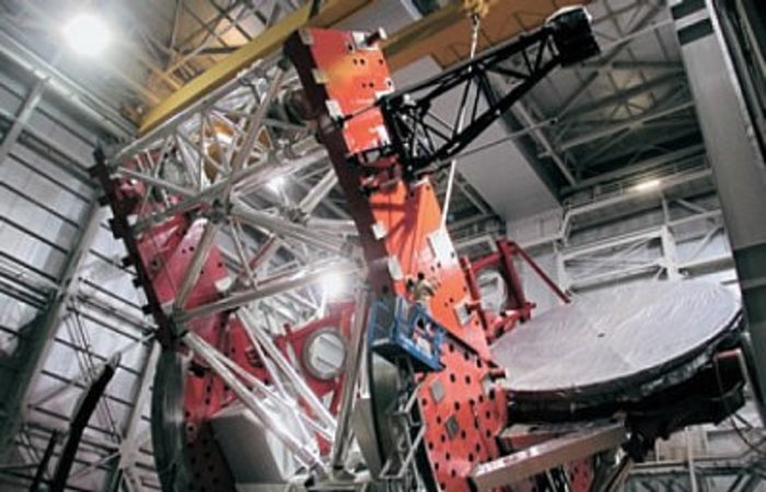 Die gewaltigen Dimensionen des LBT Riesige Metallkonstruktion in einer Halle, an die ein großer Spiegel montiert ist. Ein Arbeiter auf einer Teleskopbühne wirkt winzig.
