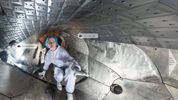 Eine Wissenschaftlerin im weißen Schutzanzug und mit Haube hockt in einem metallenen Tunnel, dessen Wände unten von wabenförmigen und oben von kassettenförmigen, durchlöcherten Platten gebildet werden.