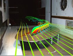 Tischartige Stahlplatte in einem kahlen Windkanal. Als Computergrafik ist eine Zugsilhouette eingefügt. Sie wird von schmalen Bändern umschlängelt, die Luftbewegungen zeigen sollen. Der Zug steht dabei schräg zur Windrichtung.