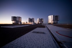 Auf der Spitze des Cerro Paranal zeigt die Fotografie das Very Large Telescope aus einiger Entfernung in der Dämmerung. Die vier Hauptteleskope, beziehungsweise die Gebäude, in denen sie sich befinden, sind dabei klar zu erkennen.