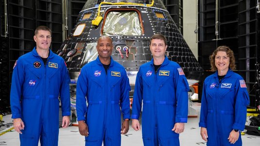 Vier Personen in blauen NASA-Fluganzügen stehen lächelnd in einer Halle. Hinter ihnen dominiert eine schwarze, kegelförmige Raumkapsel mit vielen Kacheln, Fenstern und gelben Kabeln; rundherum technische Gestelle und ein heller Boden.