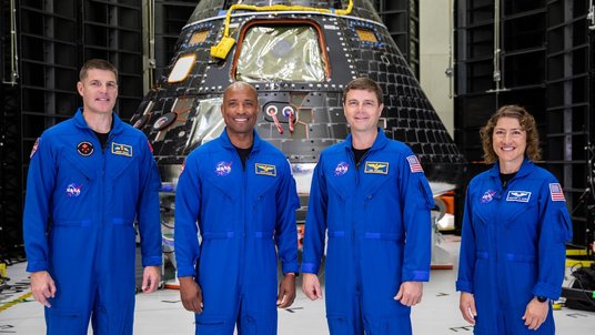 Vier Personen in blauen NASA-Fluganzügen stehen lächelnd in einer Halle. Hinter ihnen dominiert eine schwarze, kegelförmige Raumkapsel mit vielen Kacheln, Fenstern und gelben Kabeln; rundherum technische Gestelle und ein heller Boden.