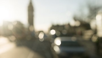 Unscharfe Stadtansicht im Gegenlicht. Im Hintergrund befindet sich die Silhouette einer Stadt mit einem markanten Kirchturm oder Glockenturm links der Bildmitte. Rechts sind verschwommene Baumkronen und Gebäudedächer abgebildet.