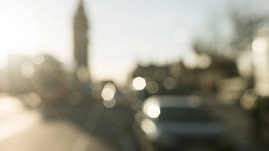 Unscharfe Stadtansicht im Gegenlicht. Im Hintergrund befindet sich die Silhouette einer Stadt mit einem markanten Kirchturm oder Glockenturm links der Bildmitte. Rechts sind verschwommene Baumkronen und Gebäudedächer abgebildet.
