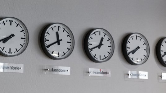 Foto. Fünf Uhren an einer Wand. Darunter stehen die Namen der Städte New York, London, Frankfurt, Hongkong und Tokyo.
