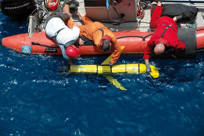 Argo-Gleiter Foto. Blick aus der Luft senkrecht nach unten auf eine Wasserfläche. Männer in einem Schlauchboot setzen ein Gerät ins Wasser ab, das einem Torpedo ähnelt. Das Gerät ist ungefähr 150 Zentimeter lang und hat kurze Flügel an der Seite und ein Steuerruder am Heck.