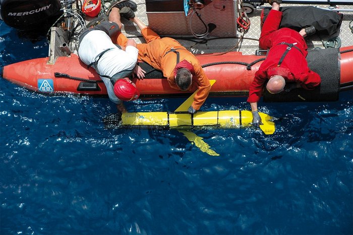 Foto. Blick aus der Luft senkrecht nach unten auf eine Wasserfläche. Männer in einem Schlauchboot setzen ein Gerät ins Wasser ab, das einem Torpedo ähnelt. Das Gerät ist ungefähr 150 Zentimeter lang und hat kurze Flügel an der Seite und ein Steuerruder am Heck.