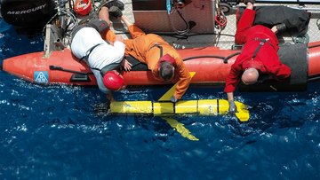 Foto. Blick aus der Luft senkrecht nach unten auf eine Wasserfläche. Männer in einem Schlauchboot setzen ein Gerät ins Wasser, das einem Torpedo ähnelt. Das Gerät ist etwa 150 Zentimeter lang und hat kurze Flügel an der Seite und ein Steuerruder am 