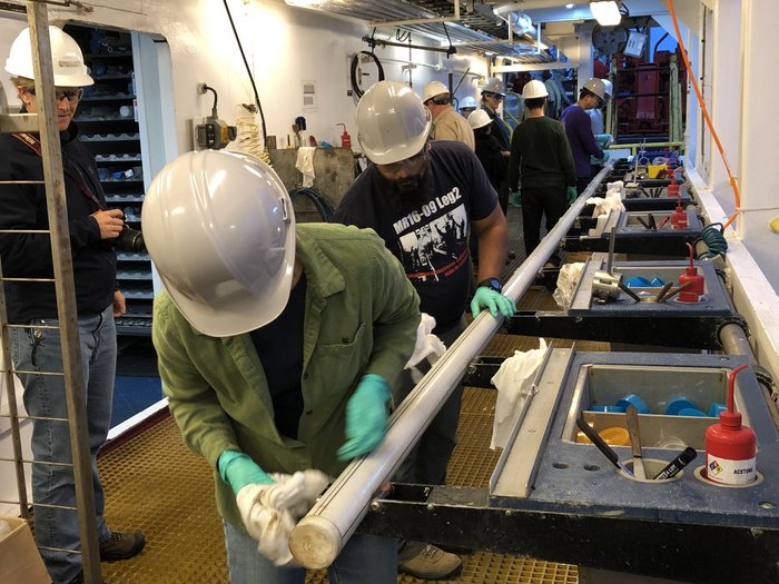 Mehrere Arbeiter mit Schutzhelm stehen an dem stangenförmigen Bohrkern und bearbeiten ihn mit kleinen Werkzeugen