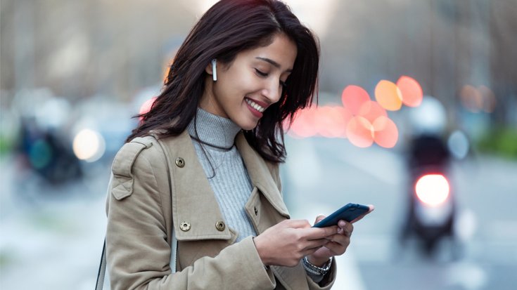 Nutzerin von Bluetooth-Kopfhörern | Bildquelle: nensuria/iStock Junge Frau, die drahtlose Kopfhörer trägt, auf ihr Smartphone schaut und lächelt. Im Hintergrund ist unscharf eine Straßenszene zu erkennen.