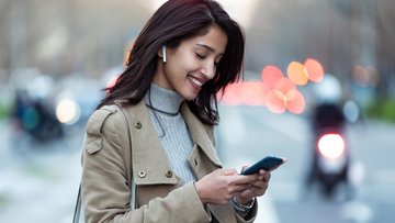 Junge Frau, die drahtlose Kopfhörer trägt, auf ihr Smartphone schaut und lächelt. Im Hintergrund ist unscharf eine Straßenszene zu erkennen.