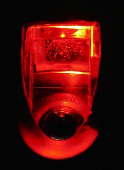 Auf schwarzem Hintergrund ist vorne der Kreis der Linse zu sehen, klares glänzendes Plastik über einem schwarzen Loch, von rotem Licht umrandet. Dahinter eine trapezförmige Struktur, von rotem Licht durchflutet, teils mit punktförmigen Einschlüssen im Kunststoff und gelblichem Licht an den Kanten.