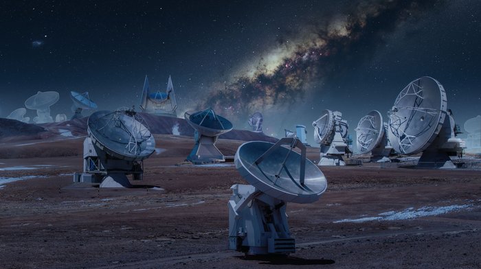 Mehrere Parabolantennen von Teleskopen in einer Wüstenlandschaft; am Nachthimmel die Milchstraße