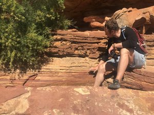 Aufnahme einer kargen Felswand mit deutlich sichtbaren, horizontal geschichteten Sedimentlagen. Rechts im Bild sitzt eine Person auf einem Felsvorsprung, mit dem Blick zur Felswand gerichtet. 
