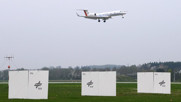 Sperrholzplatten helfen gegen Wirbelschleppen Das Bild zeigt den Rasenbereich vor einer Landebahn an einem kleinen Forschungsflughafen. Auf der Rasenfläche sind hochkant Sperrholzplatten angebracht.