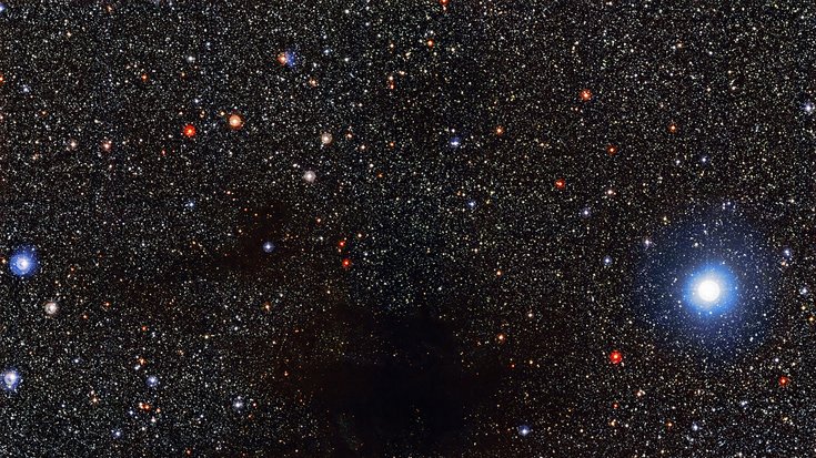 Die Dunkelwolke Lupus 4 in der Bildmitte unterbricht die dichte Anordnung heller Sternenpunkte auf dunklem Hintergrund
