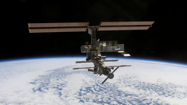 Raumstation fliegt im All, im Hintergrund ist die Erde als wolkenbedeckte Oberfläche zu sehen.