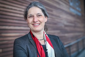 Eine Frau in einem Blazer und einem bunten Schal lächelt in die Kamera. Im Hintergrund ist unscharf eine mit Holzleisten verkleidete Wand zu erkennen.