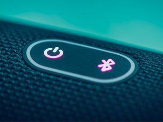 Nahaufnahme eines schwarzen, stoffüberzogenen Bluetooth-Lautsprechers mit beleuchtetem Ein-/Ausschaltknopf und leuchtendem Bluetooth-Symbol.