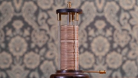 Auf dem Bild ist ein historisches wissenschaftliches Gerät abgebildet, bestehend aus einer zylindrischen Säule aus vielen abwechselnd geschichteten Metallscheiben. Vier senkrechte Glasstäbe halten die Säule und sind oben und unten in runde Halterungen eingelassen. Seitlich ragt ein metallener Hebel aus der unteren Halterung heraus.