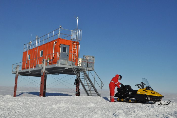 Foto. Roter Container-Bau auf Stelzen in schneeweißer Ebene, daneben ein Forscher in rotem Overall mit einem Motorschlitten.