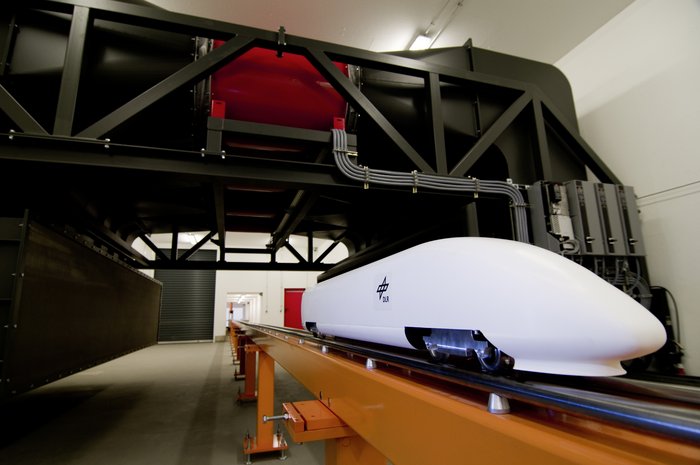 Tunnelversuchsanlage für Schnellzugmodelle Weißes Modell eines Schnellzuges auf einer langen Bahn, die durch mehrere Räume führt. Im Hintergrund die Durchbrüche durch die Wände.