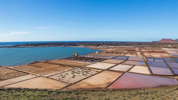 Foto: An einer Küste sind mit Steinen abgetrennte, quadratische Wasserbecken zu sehen, daneben Haufen weißen Salzes.