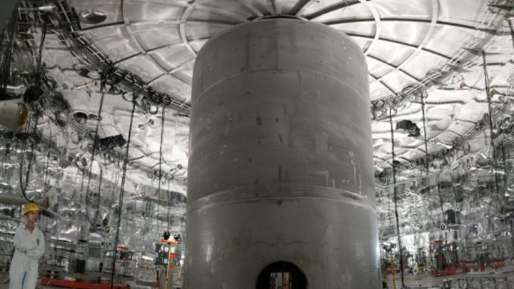 Ein Mensch steht in einer glänzend-metallenen Halle neben einem großen, stumpf-metallenen Zylinder.