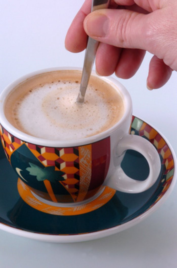 Der Klang beim Kaffeeumrühren Kaffeetasse, mit Cappuccino darin, eine Hand rührt darin mit einem Löffel.