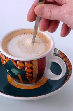 Kaffeetasse, mit Cappuccino darin, eine Hand rührt darin mit einem Löffel.