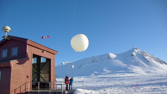 Zwei Personen vor einem roten Holzhaus mit großem, geöffnetem Tor blicken nach oben auf einen 3 Meter durchmessenden Ballon, der gerade gestartet ist. An ihm hängt eine Schuhkartongroße Box. Im Hintergrund eine schneebedeckte Landschaft mit Bergen.