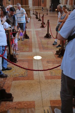 Foto des Innenraums einer Kirche: Auf dem Boden sieht man einen metallenen Streifen, auf dem ein heller Lichtkreis liegt - das Sonnenlicht; um in herum stehen Menschen und schauen sich die Linie an