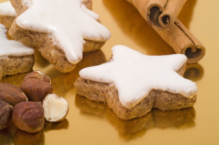 Zimtsterne Zwei fünfeckige braune Plätzchen sind mit Zuckerguss überzogen. Daneben einige Haselnüsse und Zimtstangen.