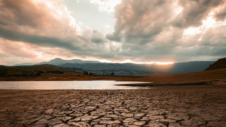 Weite Landschaft mit ausgetrocknetem, rissigem Boden im Vordergrund, einem flachen Gewässer und Bergen im Hintergrund unter dramatisch bewölktem Himmel bei Sonnenuntergang.