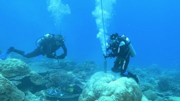 Zwei Taucher unter Wasser, einer von ihnen stellt einen metallenen Zylinder auf einen Felsen