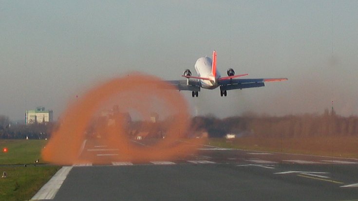 Das Bild zeigt die Rückansicht eines landenden Flugzeuges. Hinter der linken Tragfläche hat sich ein Wirbel aus Rauch gebildet. 