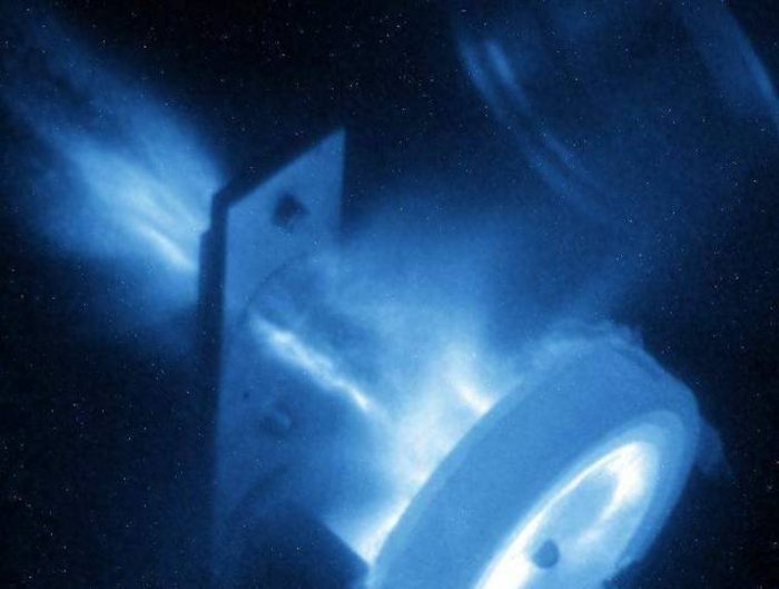 Laser beschleunigt Protonen In der Bildmitte ist eine Halterung aus Metall zu erkennen, von der in beide Richtungen filamentartige leuchtende Strukturen ausgehen. Rechts befindet sich ein ringförmiges Gebilde.