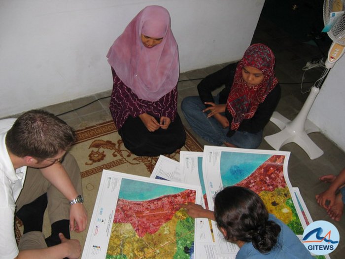 Foto. Einwohner Sumatras sitzen auf dem Boden vor Karten mit Überschwemmungsgebieten, die ihnen von Fachleuten erläutert werden.