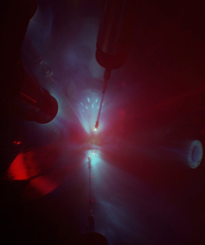 Versuchskammer mit Magnesiumoxid Blick in eine tiefe, enge Kammer, die nur schwach rot und blau erleuchtet ist. Mehrere Stangen, an deren Ende helleres Licht zu sehen ist, ragen hinunter.