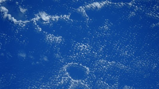 Die Meeresoberfläche aus dem All gesehen. Gleichmäßige Wolken überziehen den Himmel, außer in einer kreisförmigen, wolkenfreien Region.