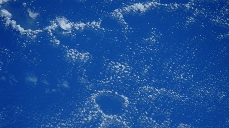 Die Meeresoberfläche aus dem All gesehen. Gleichmäßige Wolken überziehen den Himmel, außer in einer kreisförmigen, wolkenfreien Region.
